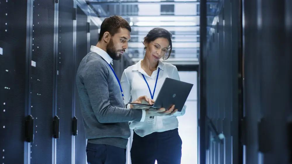 Male IT Specialist Holds Laptop and Discusses Work with Female Server Technician. They're Standing in Data Center, Rack Server Cabinet is Open.
