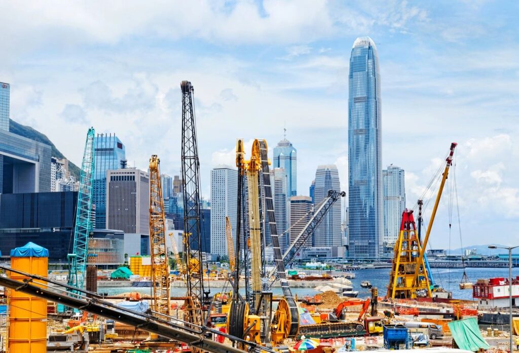 A busy port with construction cranes and machinery, set against a backdrop of modern skyscrapers and a clear blue sky. Physical and cybersecurity.