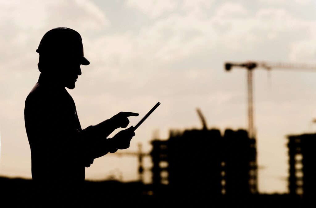 Silhouette of a worker wearing a helmet, holding a tablet and pointing, with cranes and buildings in the background.